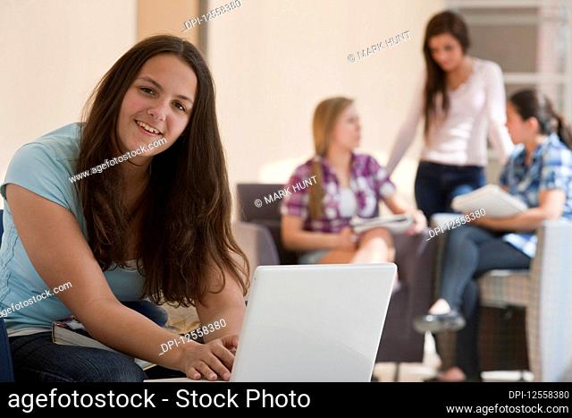 Teenage girl using laptop at school with two students looking at textbooks and talking in the background