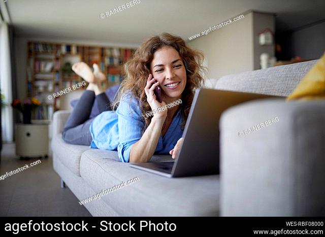 Smiling woman talking on mobile phone while lying on sofa at home
