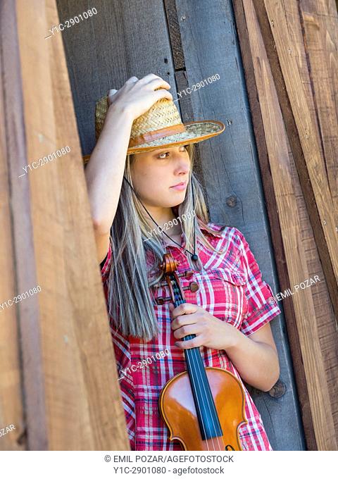 Country-girl with violin standing on door holding hat looking away in wild west environment. Croatia