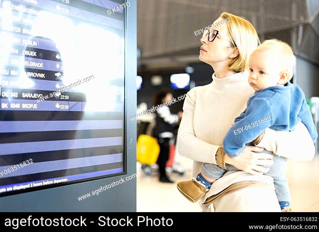 Mother traveling with child, holding his infant baby boy at airport terminal, checking flight schedule, waiting to board a plane