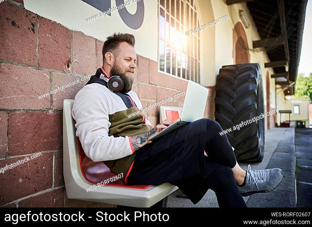 Bearded male fitness instructor using laptop while sitting on bench outside gym