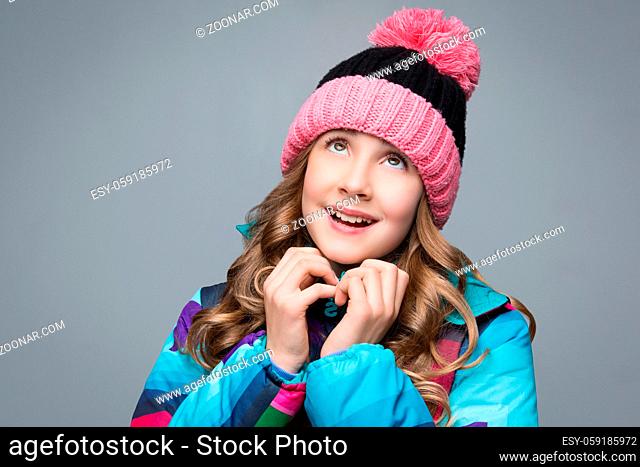 Beautiful happy teen girl with long curly hair in pink wool hat and bright warm coat looking up. Studio shot over grey background. Copy space