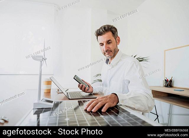 Engineer holding mobile phone analyzing solar panel sitting with laptop at desk