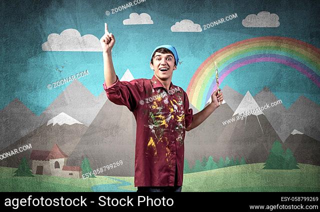 Young male artist with paintbrush. Happy painter in shirt and bandana standing on background colorful picture on wall. Summer landscape with mountains and...