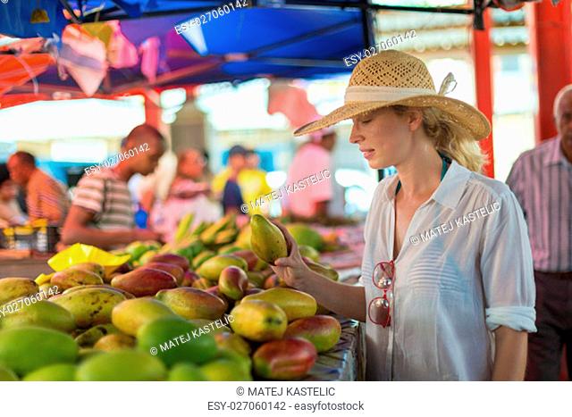 Female traveler wearing elegant colonial style white tunic and hat buying fresh tropical fruit on traditional Victoria food market on Seychelles islands