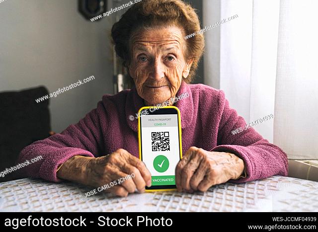 Elderly woman showing COVID-19 vaccination certificate on smart phone at home