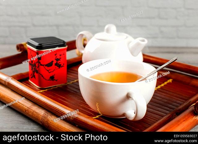 Cup of hot black tea with teapot and metal tea dose in background, served on bamboo wood tray