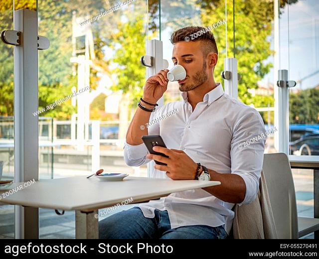 Close up of Handsome Young Man Drinking Coffee at the Shop While Looking at Cell Phone