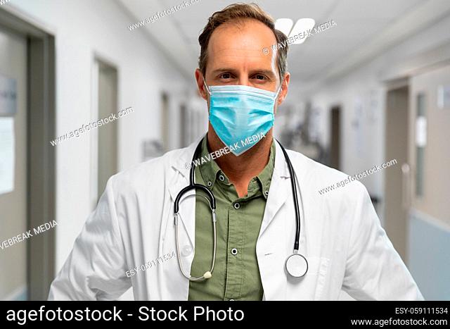 Portrait of caucasian male doctor wearing face mask standing in hospital corridor