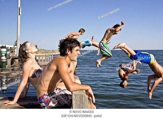 Young Couple Sitting on Edge of Pier With Group of Boys Jumping in Water