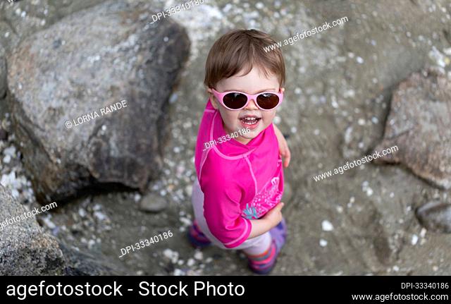 Portrait of 23 month old girl standing on a rocky beach wearing pink sunglasses looking up at the camera; North Vancouver, British Columbia, Canada