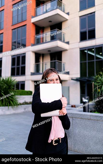 Businesswoman covering face with laptop in front of building