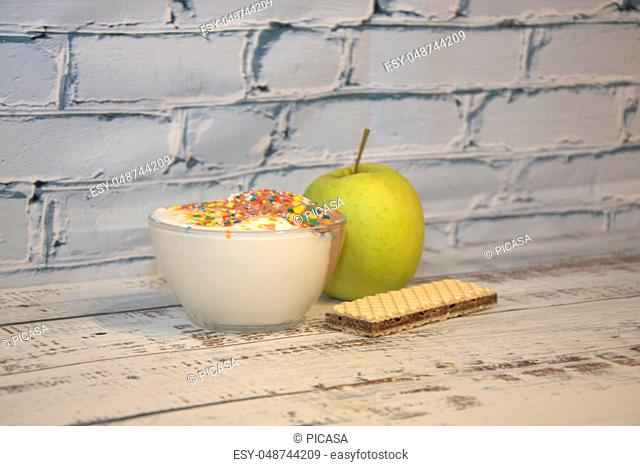 Ice cream with decor in a glass bowl along with a green apple and chocolate waffle lie on a wooden table