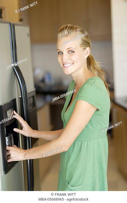 Young woman in the green dress is standing in front of fridge, is filling water glass with ice cube