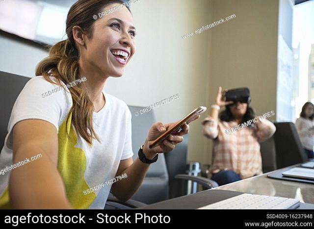 Young woman in conference room working on computer checking mobile phone, with co-worker in background wearing a Virtual reality headset