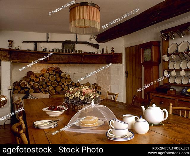 Country kitchen with a beamed ceiling, inglenook fireplace stacked with firewood, farmhouse dining table set for tea, and a ceiling light with fringed lampshade