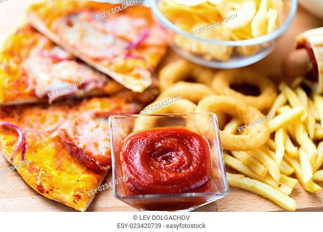 fast food and unhealthy eating concept - close up of ketchup in glass bowl over pizza, deep-fried squid rings, potato chips, peanuts and ketchup on wooden table