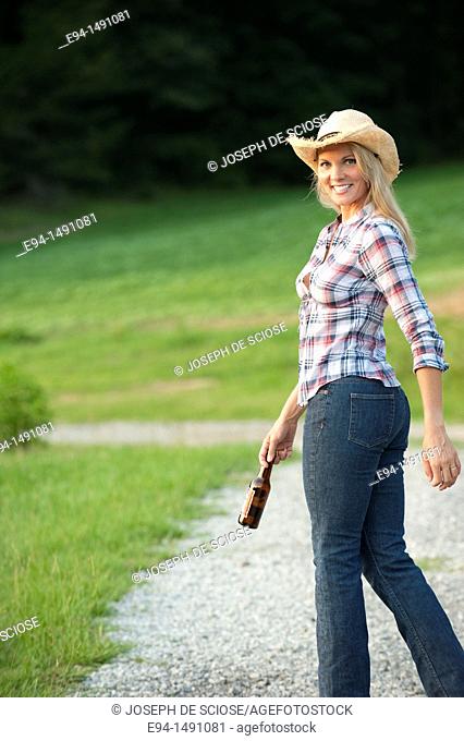 Side view of a blond woman wearing blue jeans and a straw hat waliking on a country path looking back at the camera