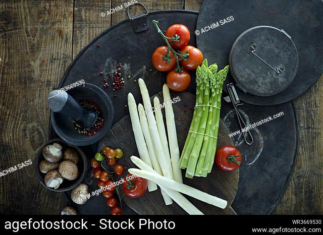 Mortar and pestle, peeler, asparagus stalks, peppercorn, tomatoes and bowl with mushrooms on rustic baking sheet
