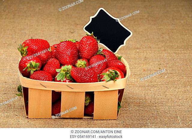 Wicker wooden basket full of red strawberries with chalk blackboard price tag sign on jute burlap canvas background, side view