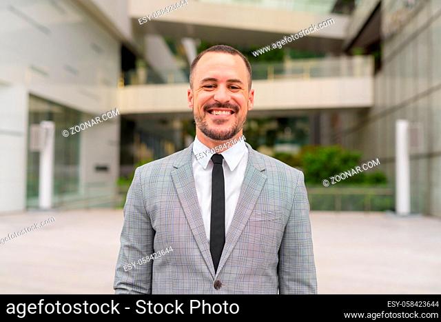 Portrait of handsome Hispanic bald bearded businessman wearing suit in the city streets outdoors