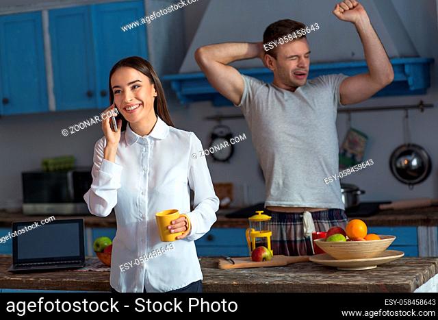 Young woman talking on phone ready to go to work. Female in business clothes taling on phone while her lazy husband yawning at background