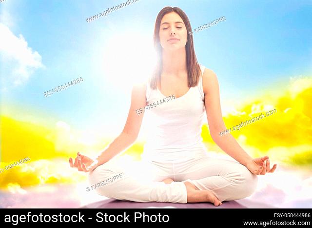 Close up full length portrait of young woman dressed in white meditating with eyes closed.Girl sitting in yoga position surrounded by clouds