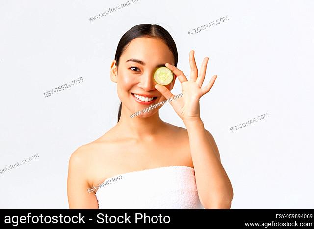Beauty, personal care, spa salon and skincare concept. Beautiful asian female in bath towel holding cucumber piece over eye and smiling