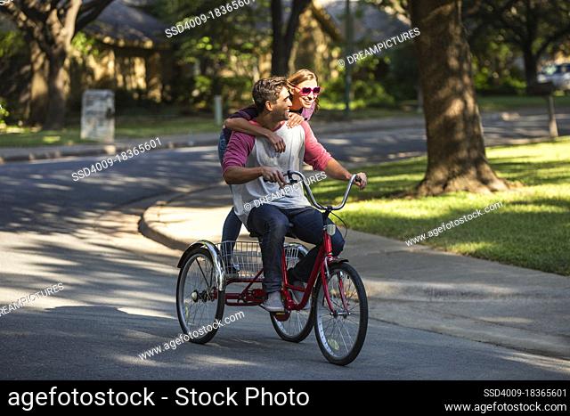 Young couple riding large tricycle through neighborhood, girl standing on back wearing heart shaped sunglasses with her arms around boyfriend