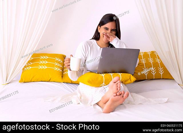 Indian woman smiling with holding a coffee mug in her hand and laptop on her lap on bed