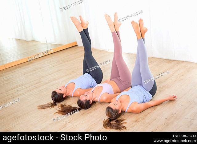 Group of three young sporty attractive women in yoga studio, lying on the floor, holding hands, legs up against white background