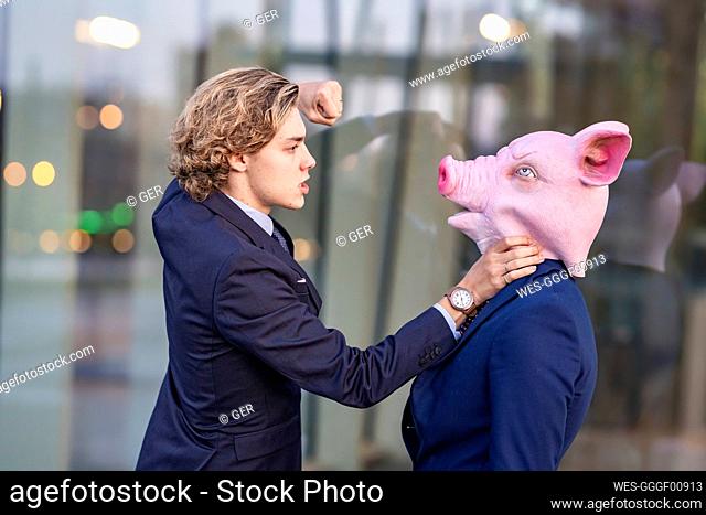 Aggressive businessman punching male coworker in pig mask by glass wall