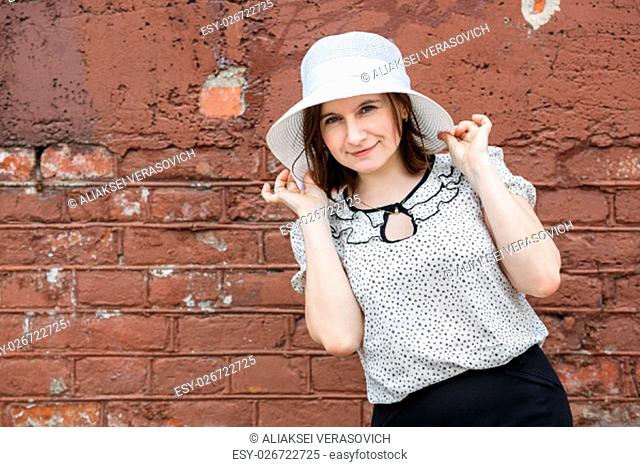 Woman in a white blouse and hat posing on a background old vintage brown brick wall. Female holds the edges of the hat by hand. Photo in retro style