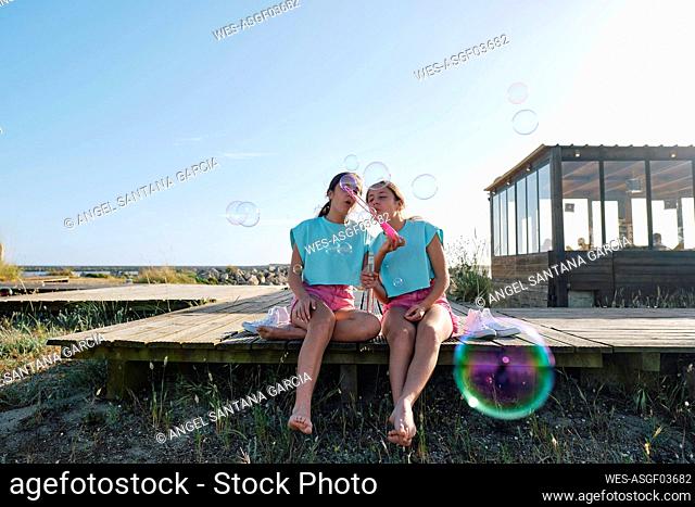 Twin sisters blowing bubbles sitting on jetty at beach