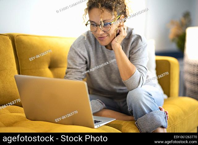 Female sitting on a yellow couch using laptop and internet connection and smile. Happy woman in indoor technology leisure activity
