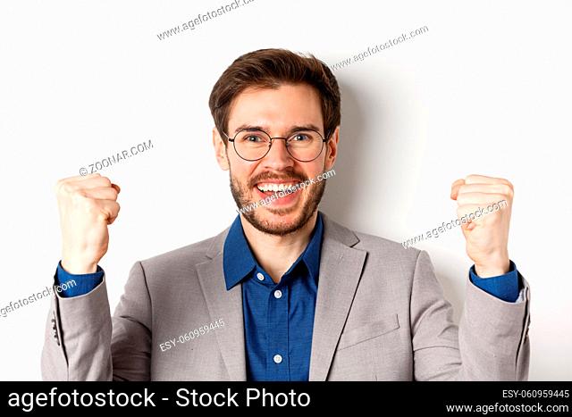 Close-up of cheerful male ceo manager in glasses and suit, saying yes and celebrating win, making fist pump, standing on white background