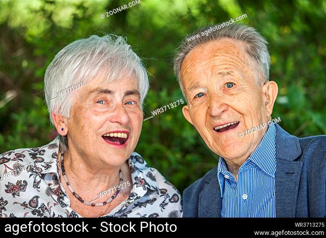 Head-and-shoulder portrait of an 80 year old retired couple in front of a blurred green background with friendly open laughter looking at the camera