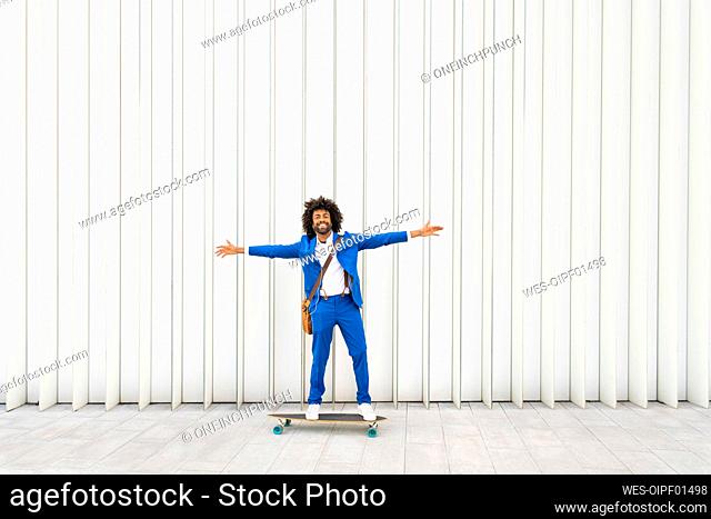 Smiling businessman standing with arms outstretched on skateboard in front of wall