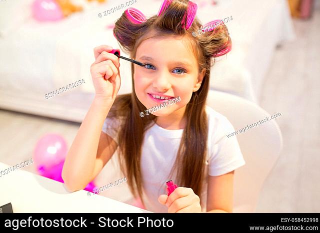 Little girl trying out her mothers cosmetics. Lovely little child with hair curlers smiling while applying some mascara onto her lashes