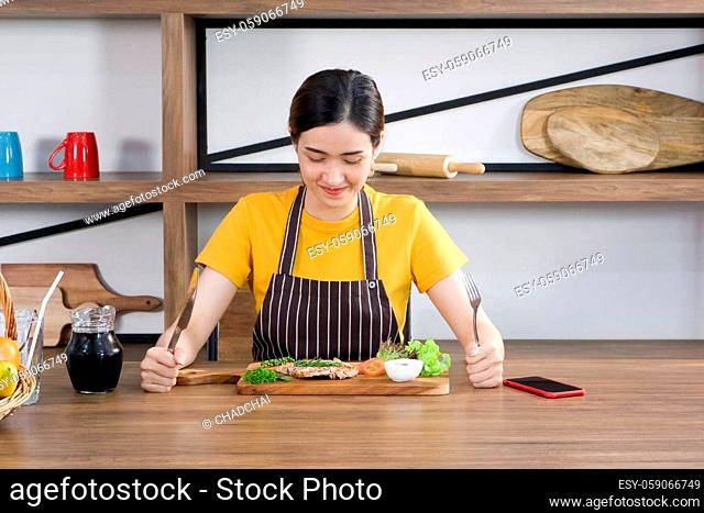 The housewife dressed in an apron, prepared a fork and knife, ready to eat a delicious steak. Morning atmosphere in a modern kitchen