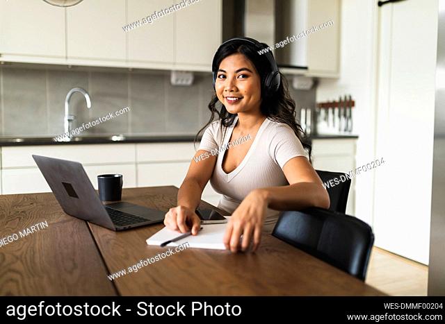 Happy freelancer wearing wireless headphones sitting at dining table in kitchen