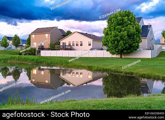 Houses trees and cloudy sky reflected on the shiny surface of a pond. The neighborhood is surrounded by a terrain covered with vivid green grasses