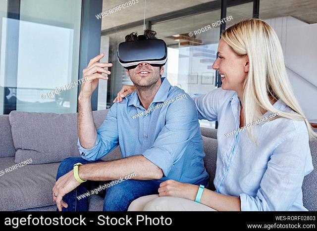 Cheerful blond woman looking at man wearing virtual reality simulator sitting in living room