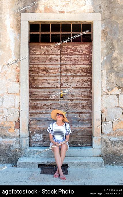 Beautiful young female tourist woman sitting and resting on vinatage wooden doorstep and textured stone wall at old Mediterranean town, smiling, holding