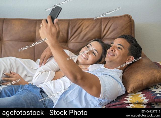 Happy romantic adult young interracial couple enjoy life at home taking selfie pictures for social media. Boy and girl together using cellular to video call...