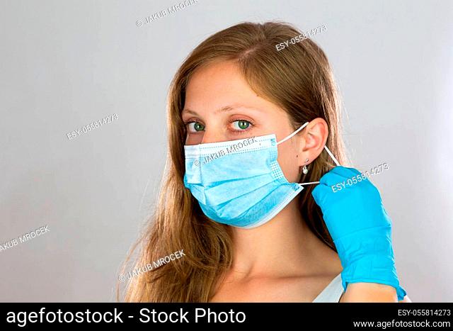 Young blonde woman putting on face mask in studio. Female model with blue gloves holding surgical mask on mouth. Lady looking to the camera protecting with...