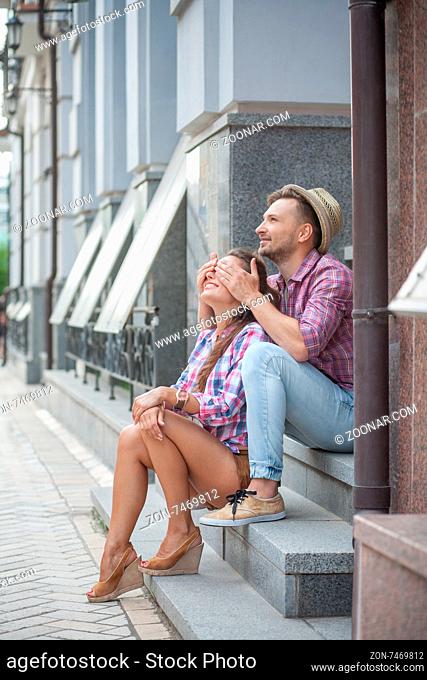 Cheerful smiling young couple having fun outside. Young man in straw hat closing eyes of his girl-friend