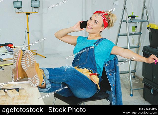 Cheerful young blond woman in jeans overalls and tool belt sitting comfortably with legs on carpenter desk talking on mobile phone with stretched out hand and...