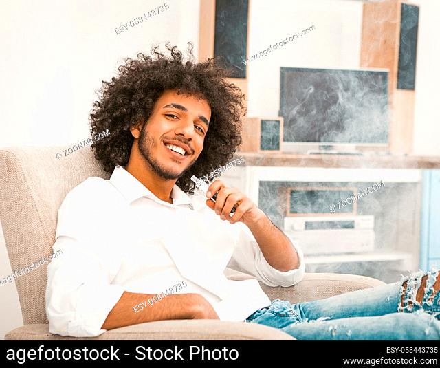 Young freelance man relaxes while smoking e-cigarette. Shaggy macho charmingly smiles while looking at the camera. Toned image