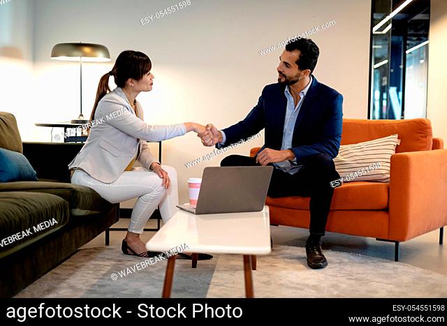 Side view of an Asian businesswoman and mixed race businessman sitting in the lobby of a modern office with a laptop computer, shaking hands and talking